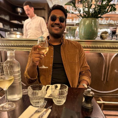 Man sitting in a restaurant booth, wearing sunglasses and holding up a glass of white wine as if cheersing to the camera.